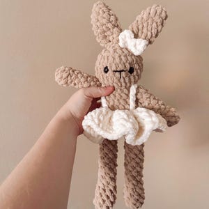 Puede incluir: Muñeca de conejo de ganchillo beige hecha a mano con tutú y lazo blancos. El conejo tiene ojos negros y boca en forma de T. La muñeca es suave y afelpada, ideal para un juguete infantil o decoración de guardería.