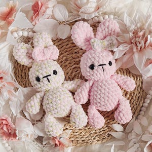 May include: Two crocheted bunny plushies, one pink and one light green with white accents, are sitting on a woven basket with a floral background.