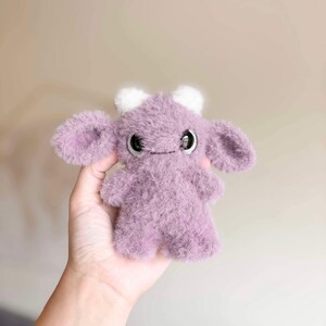 May include: A small, purple, fuzzy plush toy with large eyes and white horns. The toy is shaped like a creature with a rounded body and large ears.