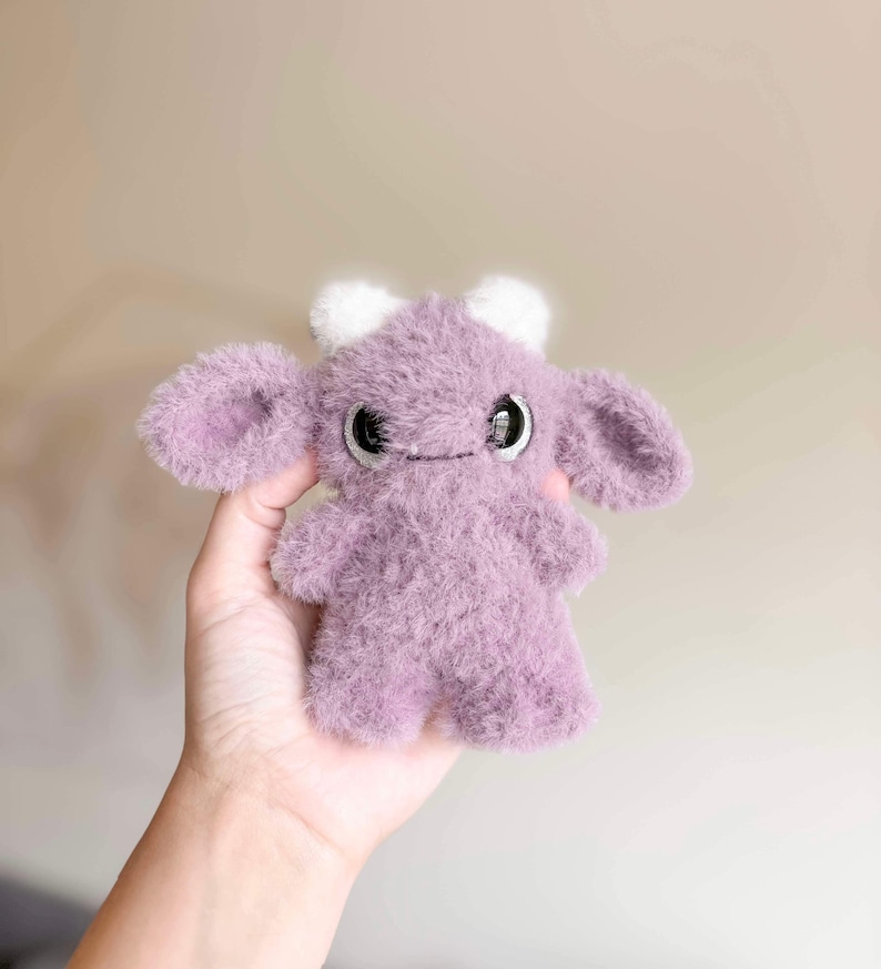 May include: A small, purple, fuzzy plush toy with large eyes and white horns. The toy is shaped like a creature with a rounded body and large ears.