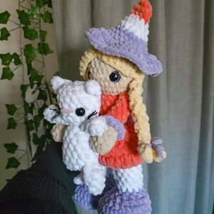May include: A handmade crochet doll with blonde braids, wearing a witch hat and dress, holding a white cat. The doll's hat is purple, white, and orange. The dress is orange and the boots are purple. The cat has black eyes and a pink nose.
