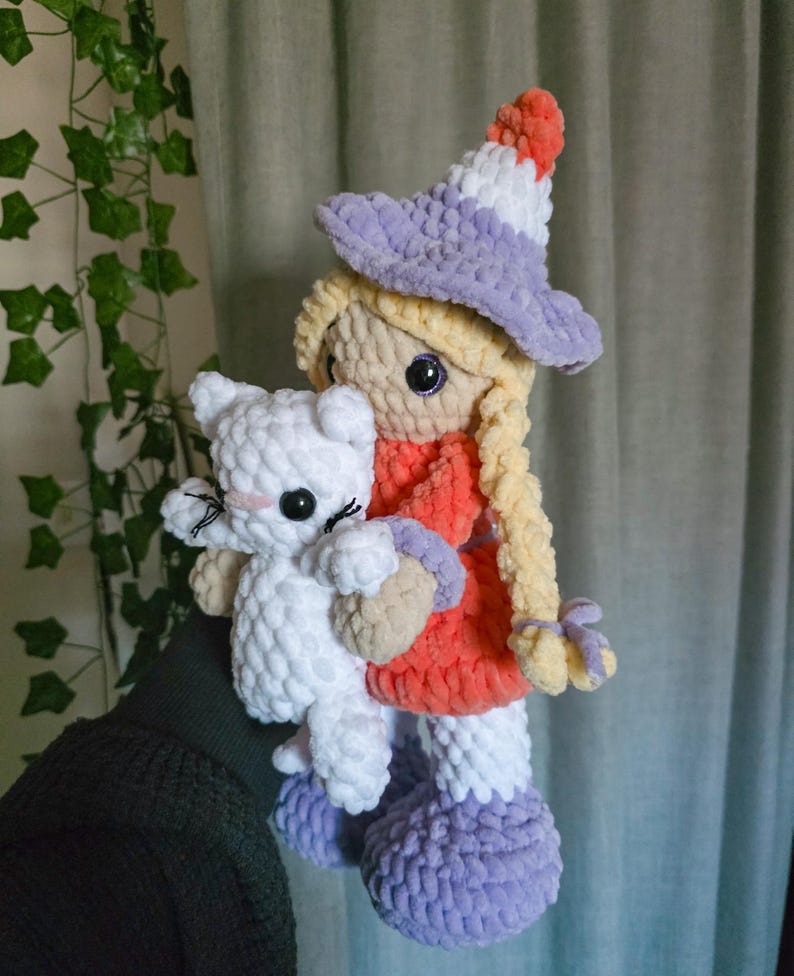 May include: A handmade crochet doll with blonde braids, wearing a witch hat and dress, holding a white cat. The doll's hat is purple, white, and orange. The dress is orange and the boots are purple. The cat has black eyes and a pink nose.
