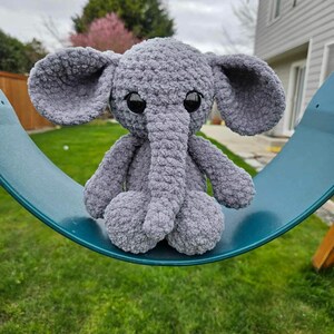 May include: A gray crocheted elephant plush toy with large black eyes sits on a teal swing set. The elephant has a long, curled trunk and floppy ears.