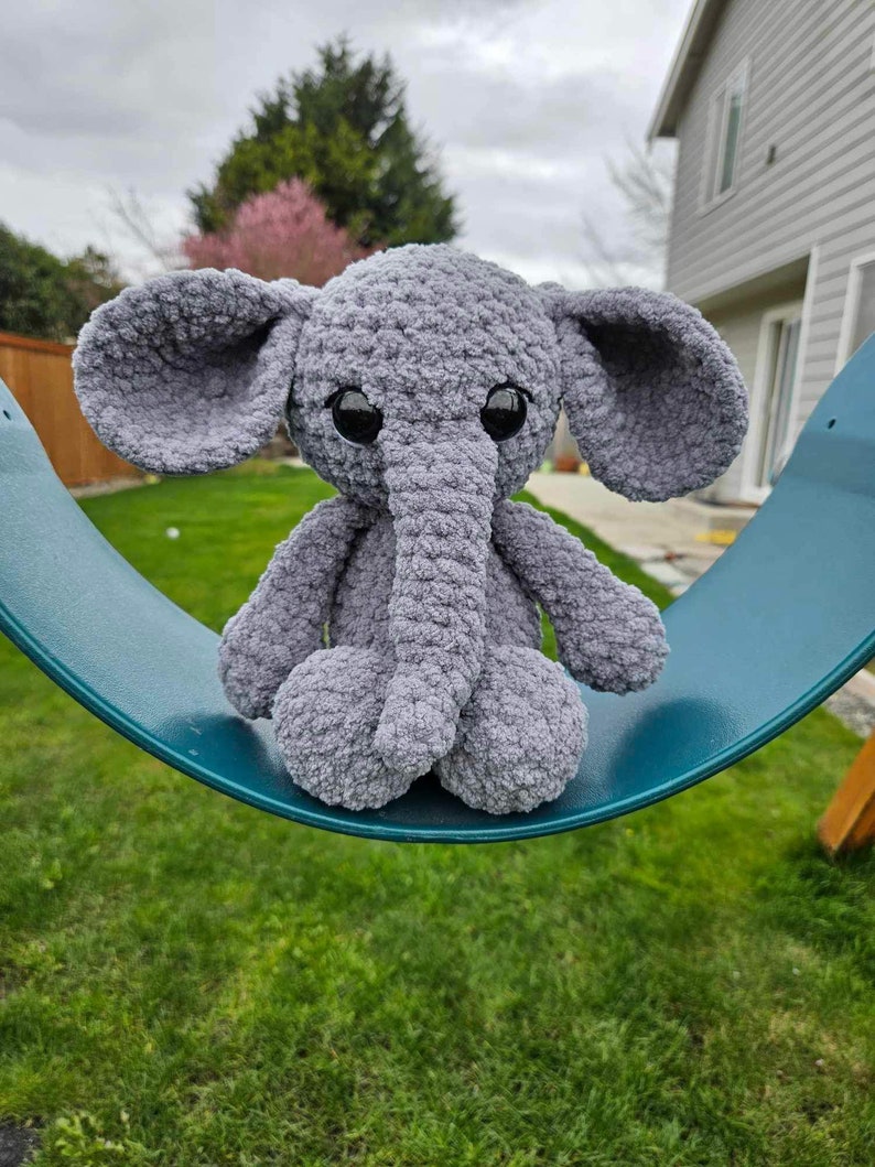 May include: A gray crocheted elephant plush toy with large black eyes sits on a teal swing set. The elephant has a long, curled trunk and floppy ears.