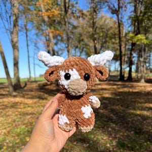 May include: A crocheted plush toy in the shape of a brown and white cow with a white face and black eyes. The toy is being held in a person's hand.