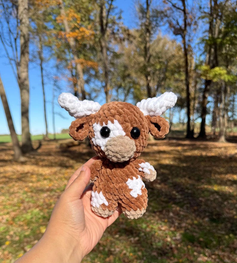 May include: A crocheted plush toy in the shape of a brown and white cow with a white face and black eyes. The toy is being held in a person's hand.