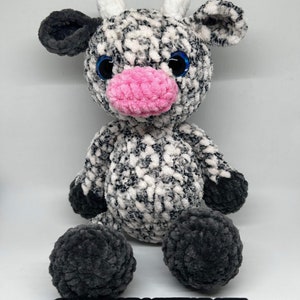 May include: A crocheted plush cow toy with black and white fur, pink nose, and blue eyes. The toy is sitting on a wooden surface with the text "@mamascrochetincrafts" written on it.