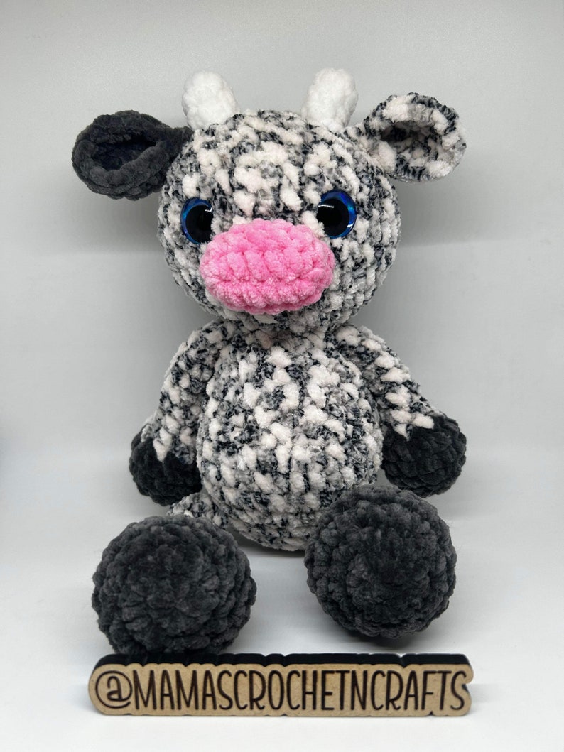 May include: A crocheted plush cow toy with black and white fur, pink nose, and blue eyes. The toy is sitting on a wooden surface with the text "@mamascrochetincrafts" written on it.