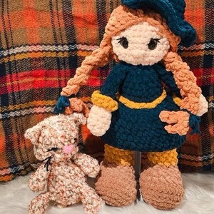 May include: A handmade crochet doll and a small cat. The doll is wearing a dark blue dress, a matching hat, and has long, braided orange hair. The cat is light brown with a pink nose. Both are made of soft, textured yarn.