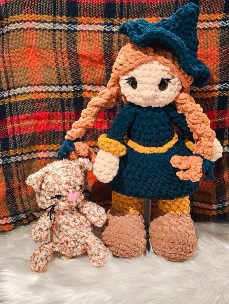 May include: A handmade crochet doll and a small cat. The doll is wearing a dark blue dress, a matching hat, and has long, braided orange hair. The cat is light brown with a pink nose. Both are made of soft, textured yarn.