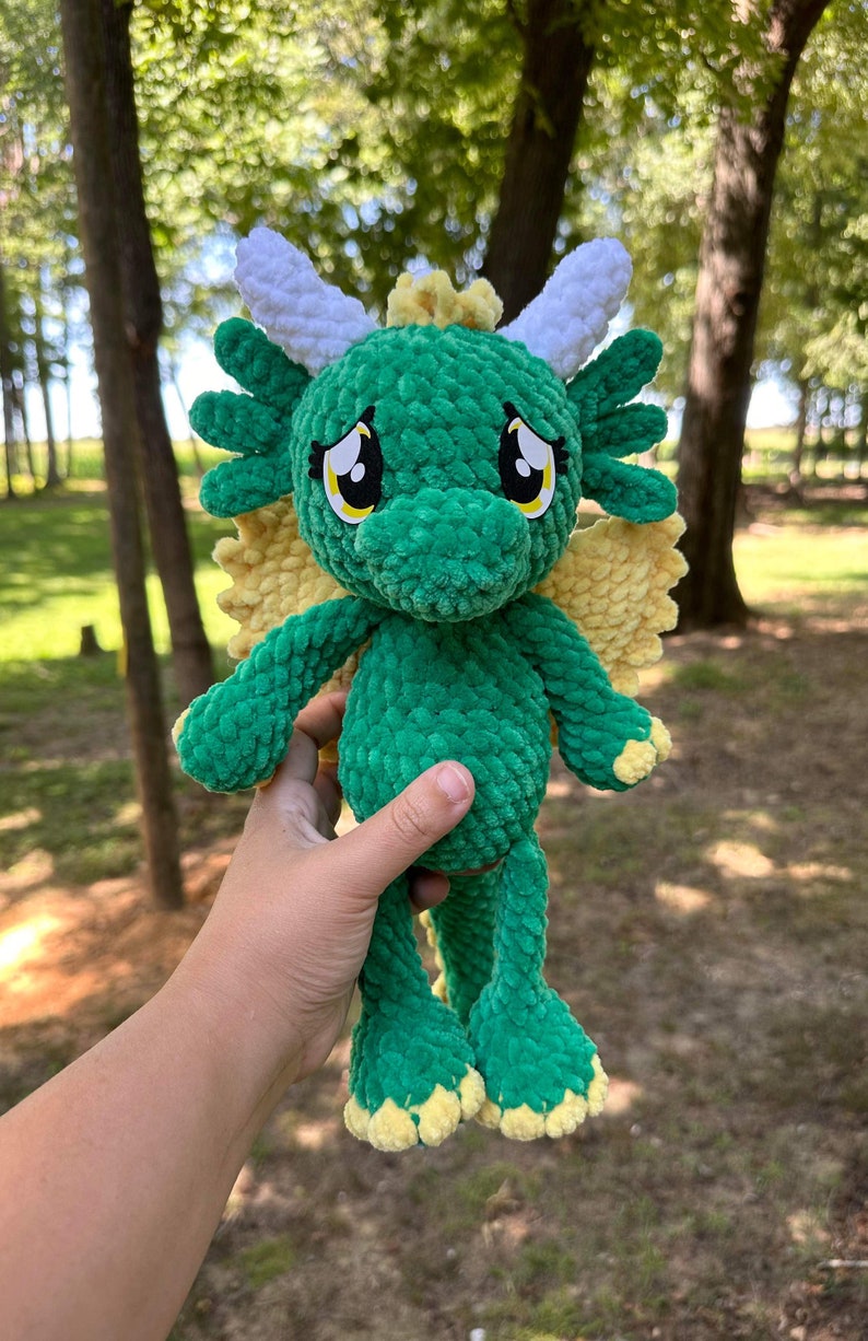 May include: A green crocheted dragon plush toy with yellow wings and white horns. The dragon has big, sad eyes and a sweet expression.