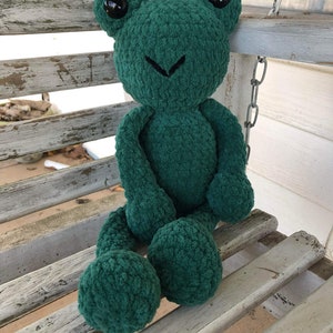 May include: A green crocheted frog plush toy with black eyes sitting on a white wooden bench.