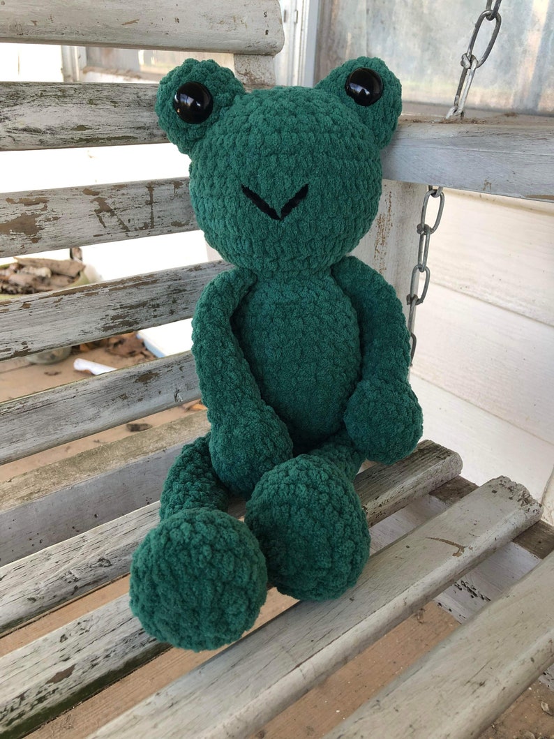 May include: A green crocheted frog plush toy with black eyes sitting on a white wooden bench.