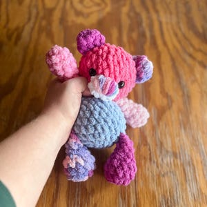 May include: A handmade crochet bear in shades of pink, purple, and blue. The bear has a textured, plush appearance with small black eyes and a multi-colored nose. The bear's body is a light blue color, with purple and pink accents on the arms and legs.