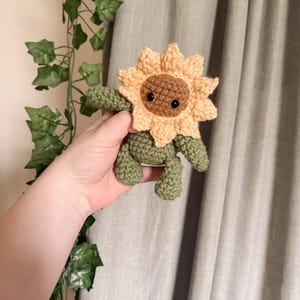 K&ouml;nnte beinhalten: Ein geh&auml;keltes Pl&uuml;sch-Sonnenblumenspielzeug mit braunem Gesicht und schwarzen Augen. Die Sonnenblume ist gelb und orange mit gr&uuml;nen Bl&auml;ttern.
