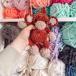 Op de afbeelding: Een handgemaakt, pluche aap-speelgoed in tinten roest, beige en bruin. De aap heeft grote, ronde oren, een klein gezicht en korte ledematen. De achtergrond toont planken gevuld met kleurrijk garen.