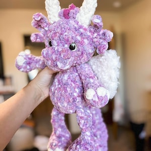 May include: A purple and white crocheted dragon plush toy with a long tail and white wings. The dragon has a pink flower on its head and large, brown eyes.