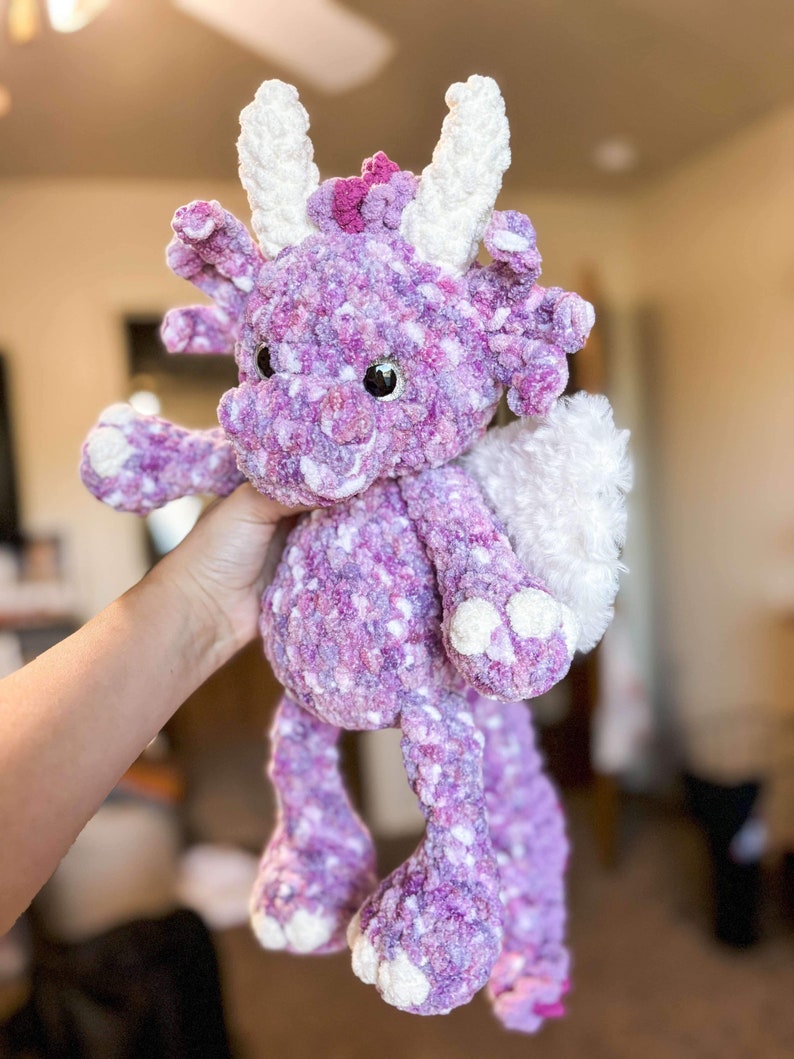 May include: A purple and white crocheted dragon plush toy with a long tail and white wings. The dragon has a pink flower on its head and large, brown eyes.