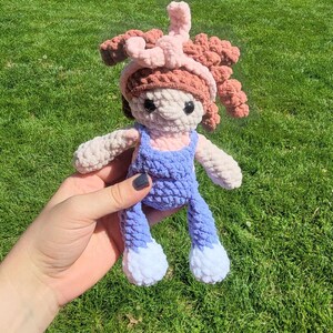 May include: A crocheted doll with pink hair, a pink headband, and a blue and white outfit. The doll is being held by a hand.