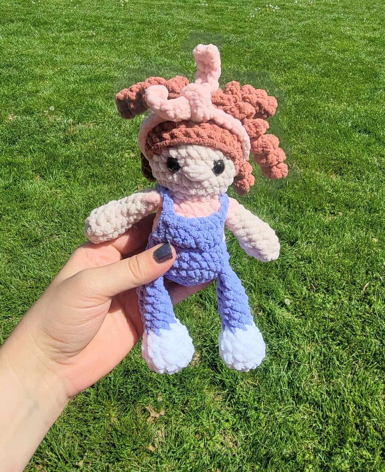 May include: A crocheted doll with pink hair, a pink headband, and a blue and white outfit. The doll is being held by a hand.