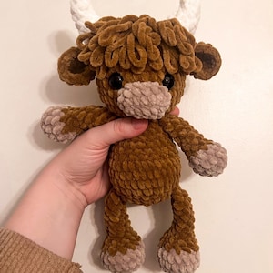 May include: A brown and white crocheted stuffed highland cow toy with a fluffy texture. The toy has a brown body, white horns, and a brown and white face.