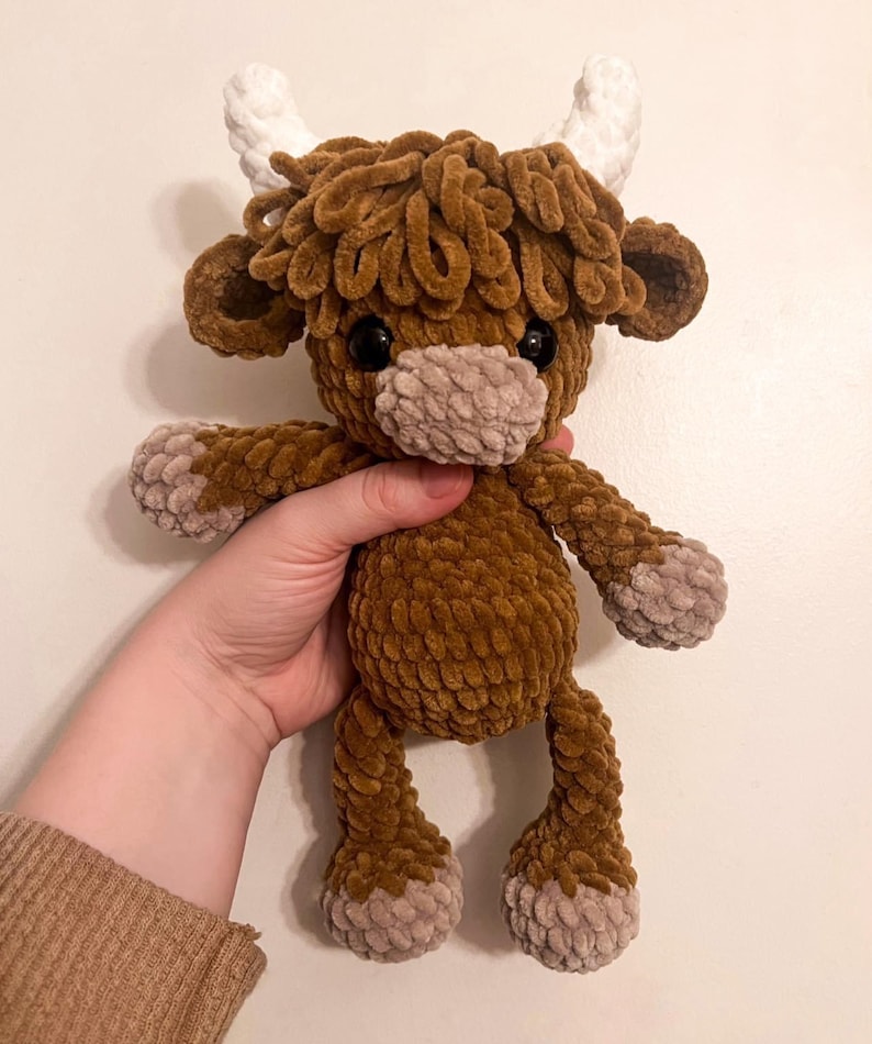 May include: A brown and white crocheted stuffed highland cow toy with a fluffy texture. The toy has a brown body, white horns, and a brown and white face.