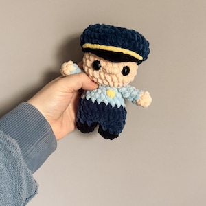 May include: A handmade crochet doll in the shape of a police officer. The doll is wearing a navy blue hat with a yellow stripe, a light blue shirt, and navy blue pants. The doll has black eyes and a yellow heart on the chest.