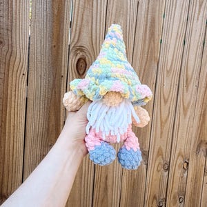 Può includere: Una bambola gnomo all'uncinetto dai colori pastello con una lunga barba bianca, naso e braccia color pesca e piedi blu. Lo gnomo indossa un cappello a balze nei toni del giallo, rosa e blu. La bambola è tenuta davanti a una staccionata di legno.