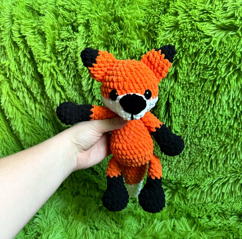 May include: A crocheted orange fox plush toy with black legs and a white face. The toy is sitting on a green fuzzy surface.
