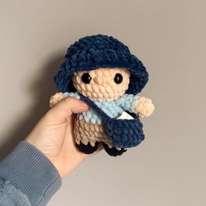 Op de afbeelding: Een gehaakt pluche speelgoed in de vorm van een persoon die een blauwe hoed draagt en een blauwe tas draagt. Het speelgoed is gemaakt met verschillende garenkleuren, waaronder blauw, beige en lichtblauw.