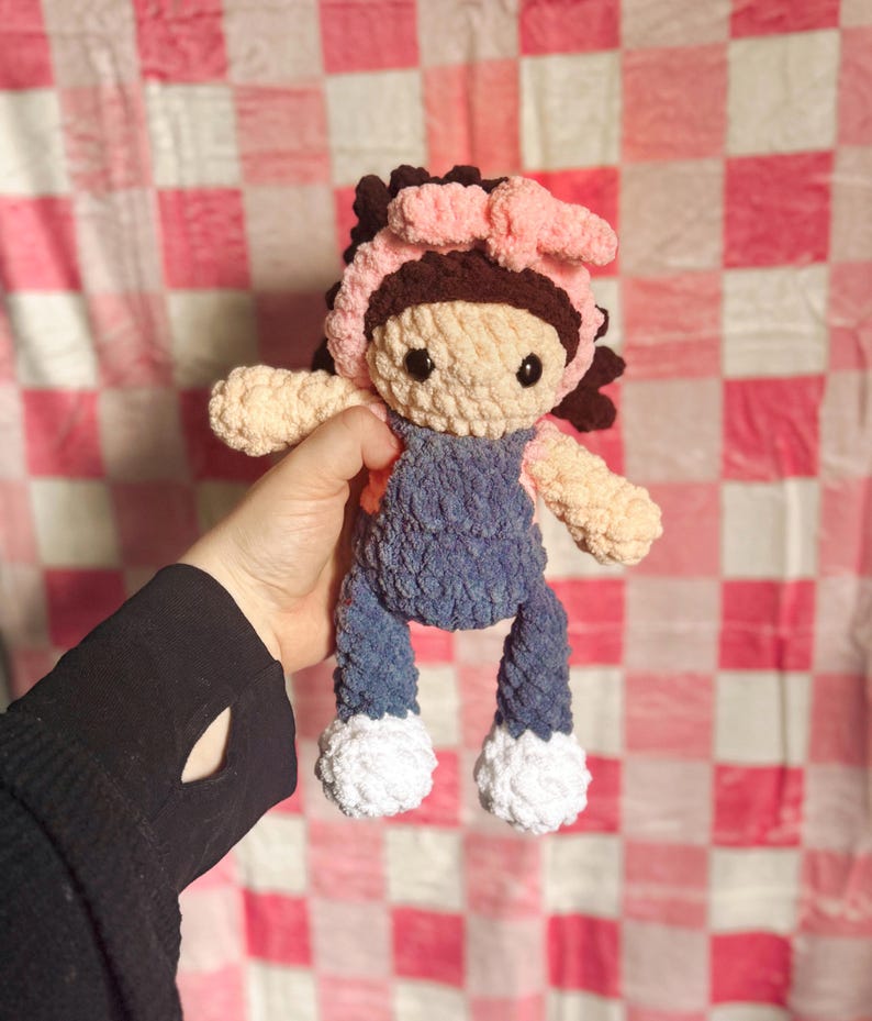 May include: A crocheted doll with brown hair, a pink bow, and a blue denim overall. The doll has white shoes and is holding a pink bow.