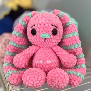 May include: A pink crocheted bunny with green striped ears. The bunny has black eyes and a green nose. The bunny is sitting on a white metal shelf. The text "@madelinemaeco" is visible on the bunny's chest.