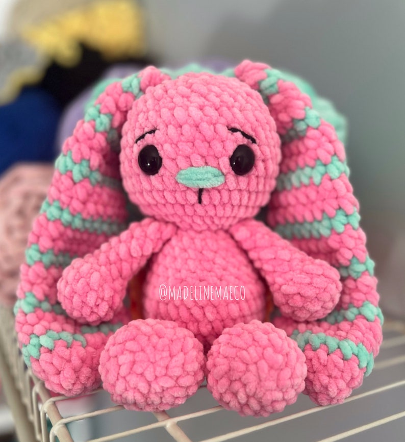 May include: A pink crocheted bunny with green striped ears. The bunny has black eyes and a green nose. The bunny is sitting on a white metal shelf. The text "@madelinemaeco" is visible on the bunny's chest.