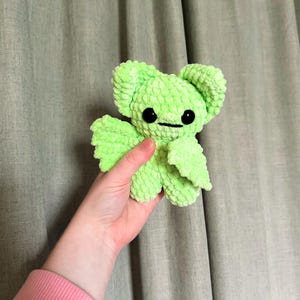 Pode incluir: Um brinquedo de pelúcia de morcego de crochê verde limão com olhos pretos. O brinquedo está sendo segurado por uma mão.