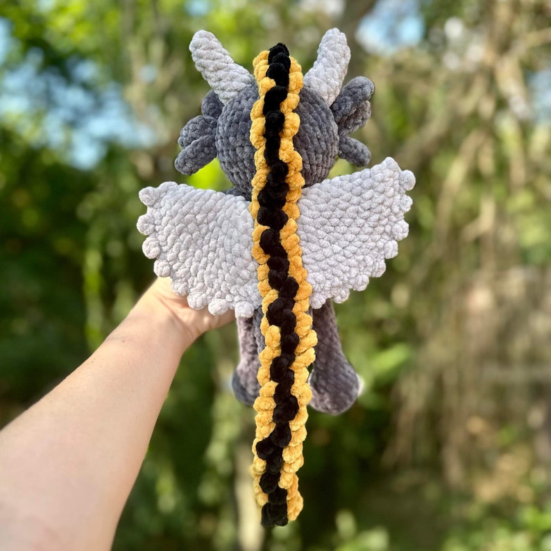 May include: A gray crocheted dragon with a long, black and yellow tail. The dragon has wings and a textured, fluffy body.