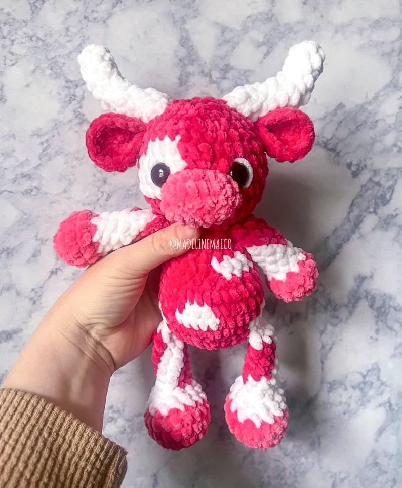 May include: A handmade plush cow toy in shades of pink and white. The cow has large, black button eyes, white horns, and a soft, textured appearance. The toy is held by a person, showcasing its size and detail. The overall theme is a cute, handcrafted animal.