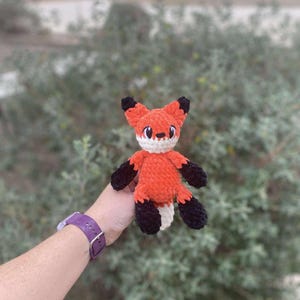 May include: A crocheted fox toy in orange and black, with a white belly and tail. The fox has black ears and paws, and a white face with black eyes. The toy is held against a blurred green backdrop.