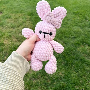 May include: A handmade pink crochet bunny with a bow. The bunny has a light pink body, a darker pink bow, and black eyes and mouth. The plush toy is soft and cuddly, perfect for a child's toy or nursery decor.