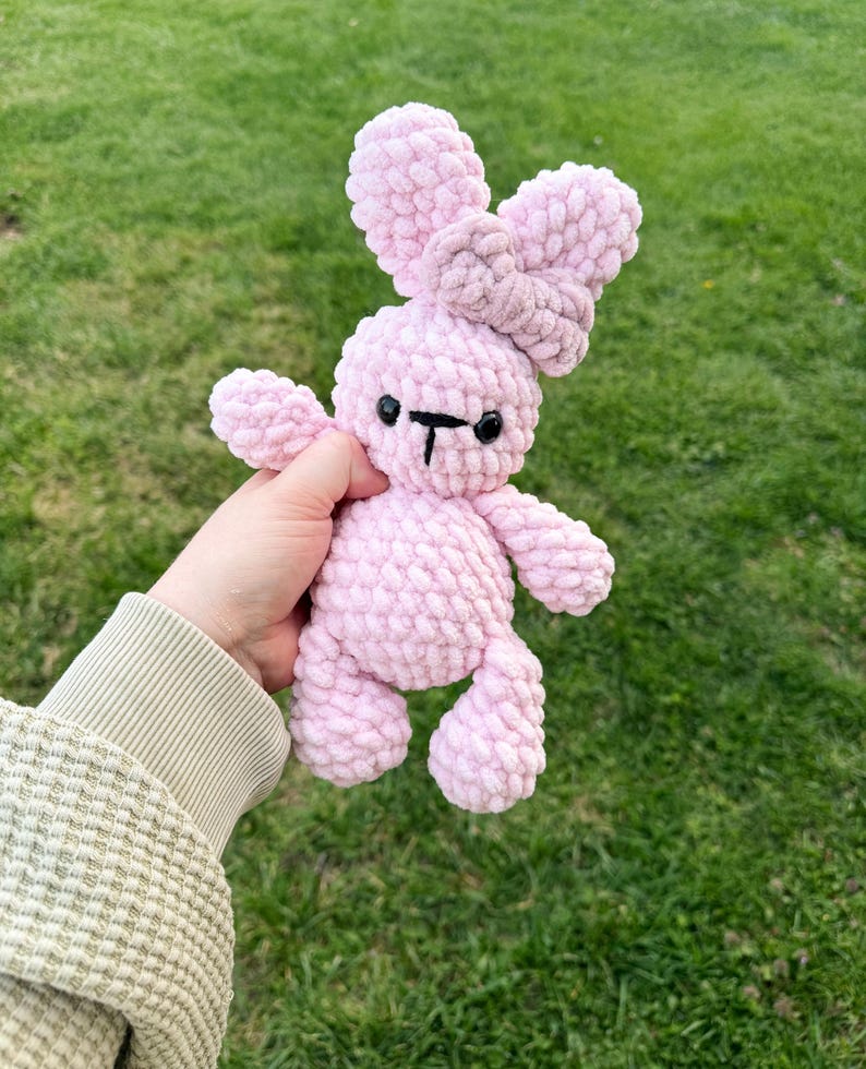 May include: A handmade pink crochet bunny with a bow. The bunny has a light pink body, a darker pink bow, and black eyes and mouth. The plush toy is soft and cuddly, perfect for a child's toy or nursery decor.
