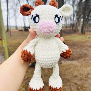 May include: A white crocheted cow plush toy with brown ears and hooves. The toy has big blue eyes and a pink nose.