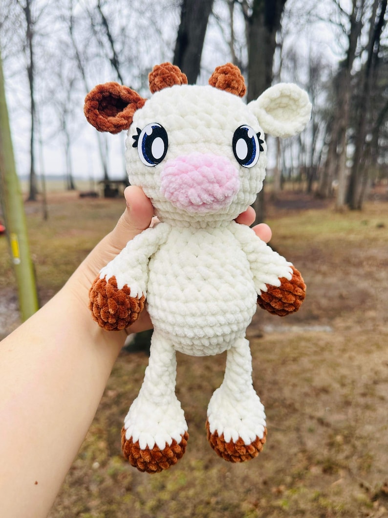 May include: A white crocheted cow plush toy with brown ears and hooves. The toy has big blue eyes and a pink nose.