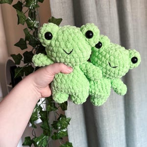 May include: Two crocheted green frog plushies with black eyes. The frogs are holding each other.