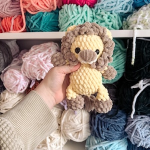 Op de afbeelding: Een handgemaakt, pluche leeuwenspeelgoed in geel- en bruintinten. De leeuw heeft een pluizige manen, kleine zwarte ogen en wordt vastgehouden voor een plank vol kleurrijke garenbollen. Het garen is in verschillende kleuren, waaronder roze, blauw en groen.