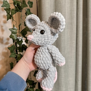 May include: A gray crocheted mouse plush toy with pink accents on the ears and feet. The toy is being held by a hand.