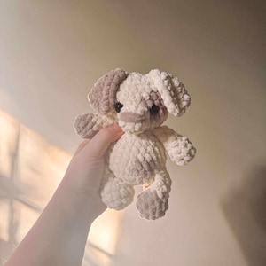 Puede incluir: Un juguete de perro de ganchillo hecho a mano en crema y marrón claro. El juguete de peluche tiene orejas caídas, un hocico pequeño y cuatro patas. El perro está en la mano de una persona.