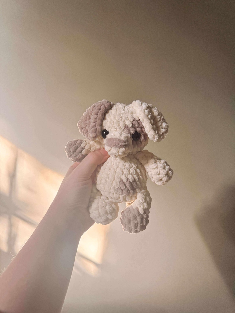 K&ouml;nnte beinhalten: Ein handgefertigtes H&auml;kelhundespielzeug in Creme und Hellbraun. Das Pl&uuml;schtier hat Schlappohren, eine kleine Schnauze und vier Beine. Der Hund wird in der Hand einer Person gehalten.