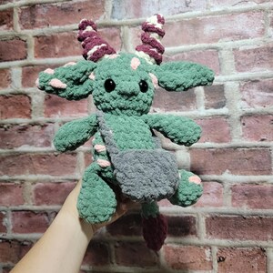 Peut inclure: Une cr&eacute;ature verte au crochet faite &agrave; la main avec des yeux noirs, des taches roses et des cornes bordeaux. Il a une sacoche grise et une queue bordeaux. La peluche est tenue contre un mur de briques.