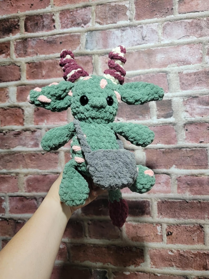 Peut inclure: Une cr&eacute;ature verte au crochet faite &agrave; la main avec des yeux noirs, des taches roses et des cornes bordeaux. Il a une sacoche grise et une queue bordeaux. La peluche est tenue contre un mur de briques.