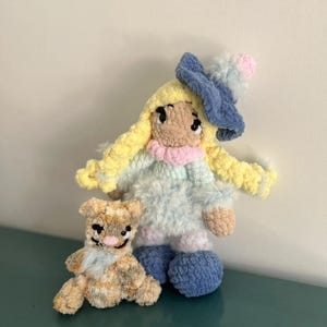 May include: Two handmade crochet dolls. One is a doll with yellow hair, a blue hat, and a light blue dress. The other is a small brown and tan teddy bear. Both dolls are made of soft yarn and have embroidered details.