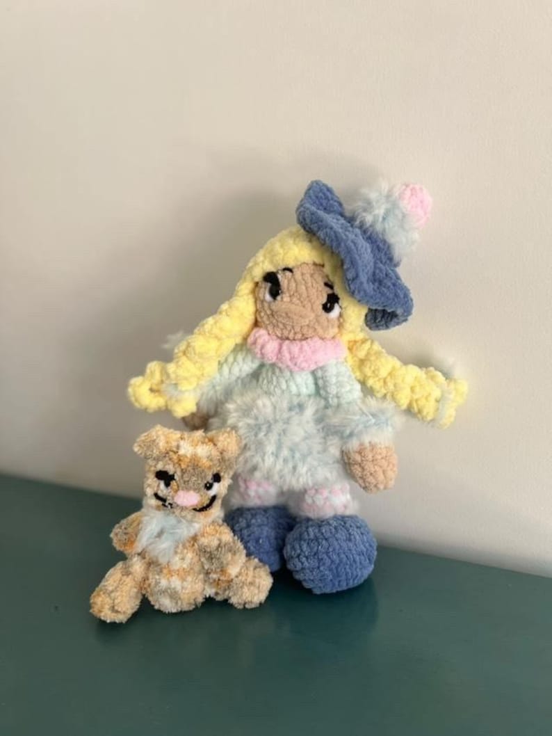 May include: Two handmade crochet dolls. One is a doll with yellow hair, a blue hat, and a light blue dress. The other is a small brown and tan teddy bear. Both dolls are made of soft yarn and have embroidered details.